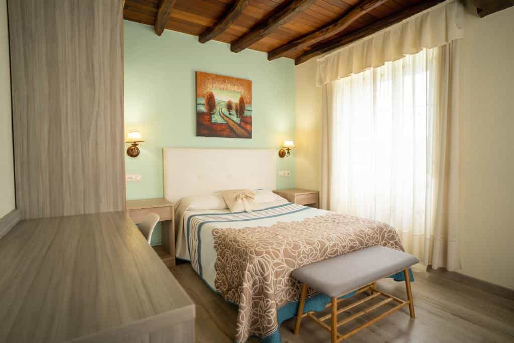 Bedroom with double bed, wooden ceiling beams, light green walls, and bright window.