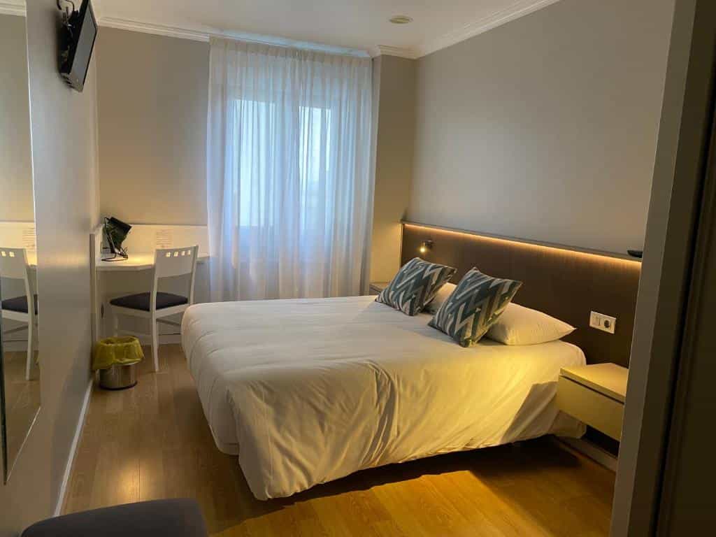 Hotel room interior with large bed, desk, chair, and sheer white curtains over a window.
