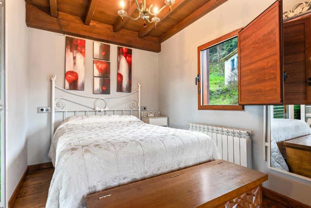 Bedroom with white metal bed, wooden ceiling beams, and view of green hillside outside.