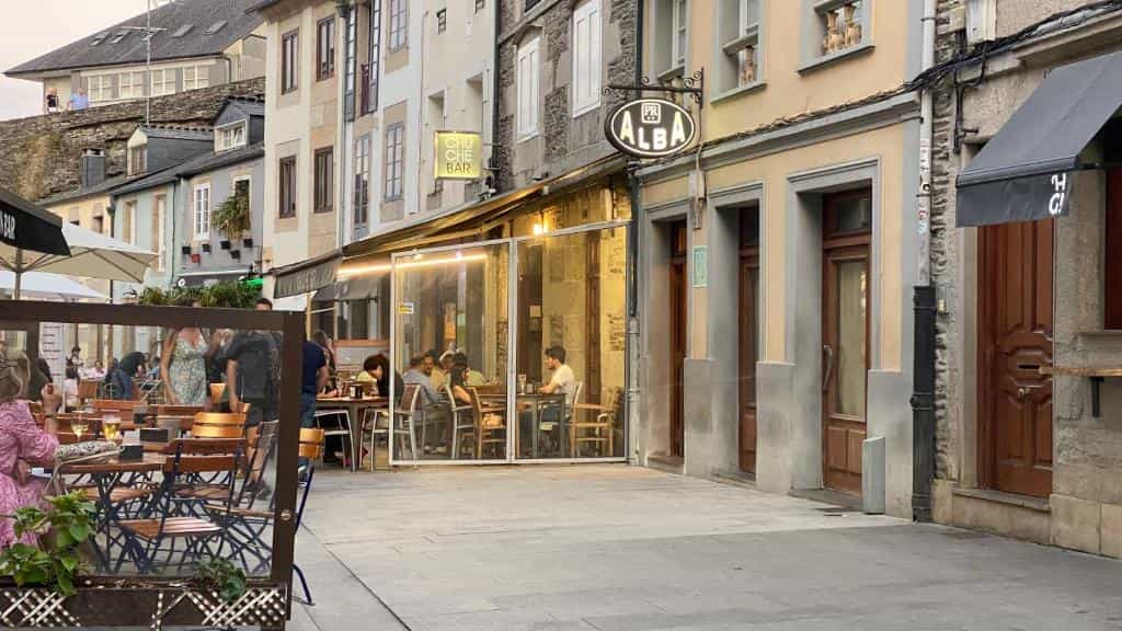 Outdoor dining patio next to Hostal Alba in Lugo with people seated at tables on a street.