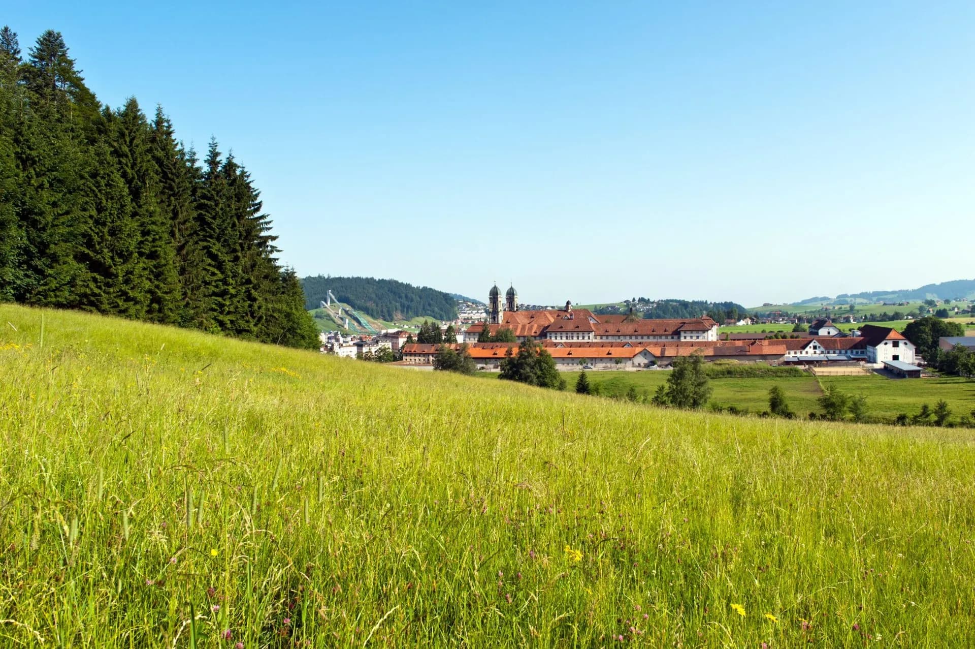 Green meadow overlooking a town with a large abbey and ski jump in the Swiss countryside.