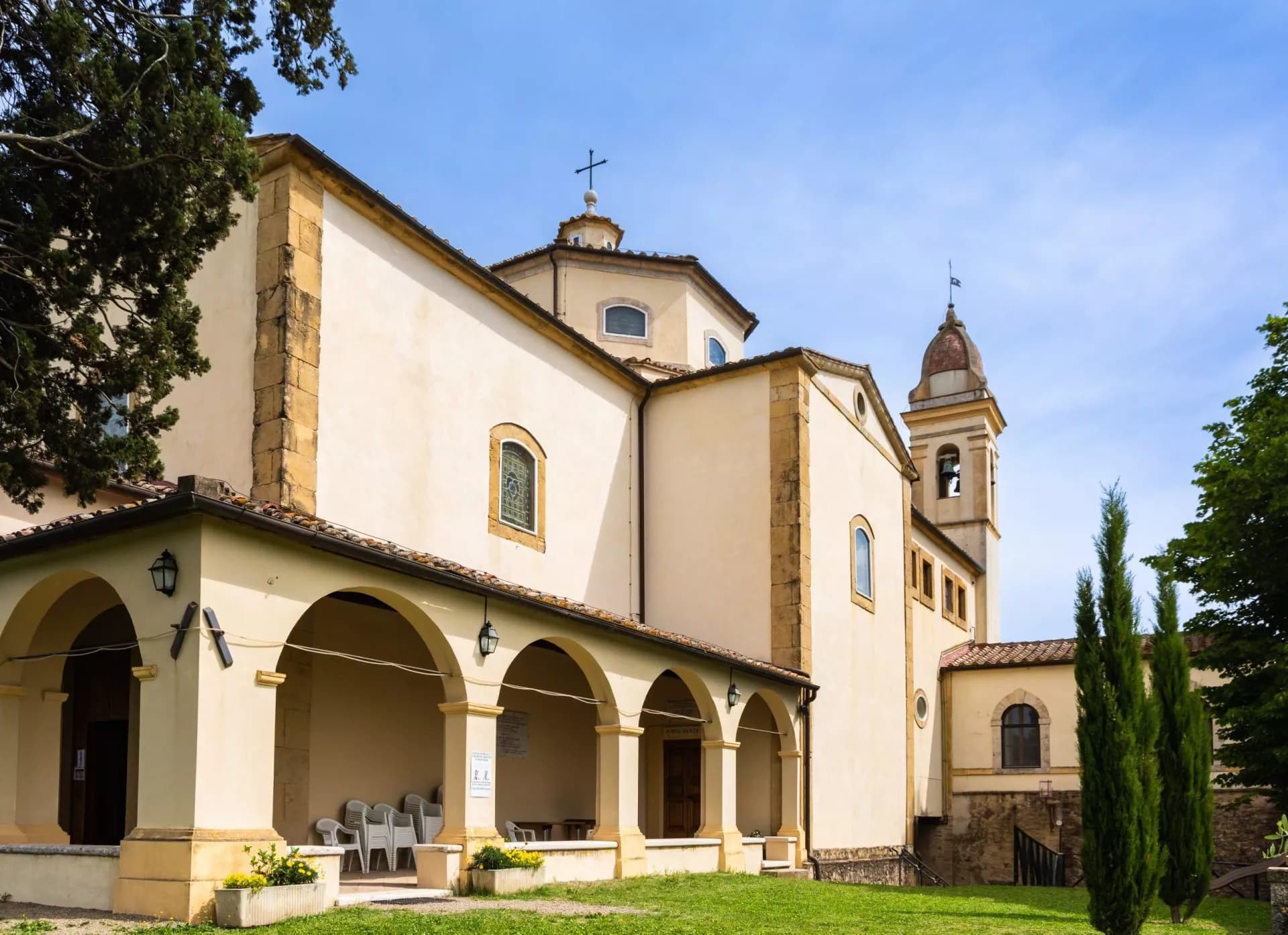 Cream-colored church with arched portico and bell tower on sunny day with cypress trees.