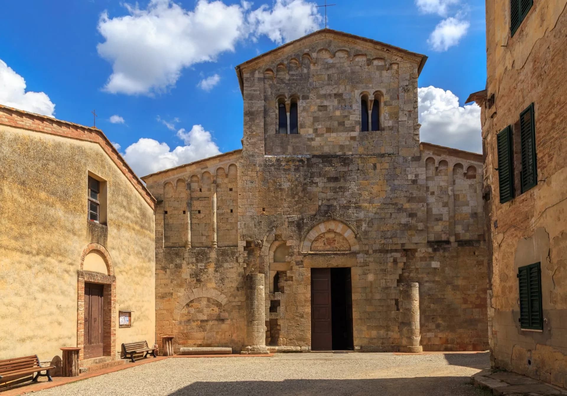 Stone church facade in Abbadia a Isola square under a bright blue, cloudy sky