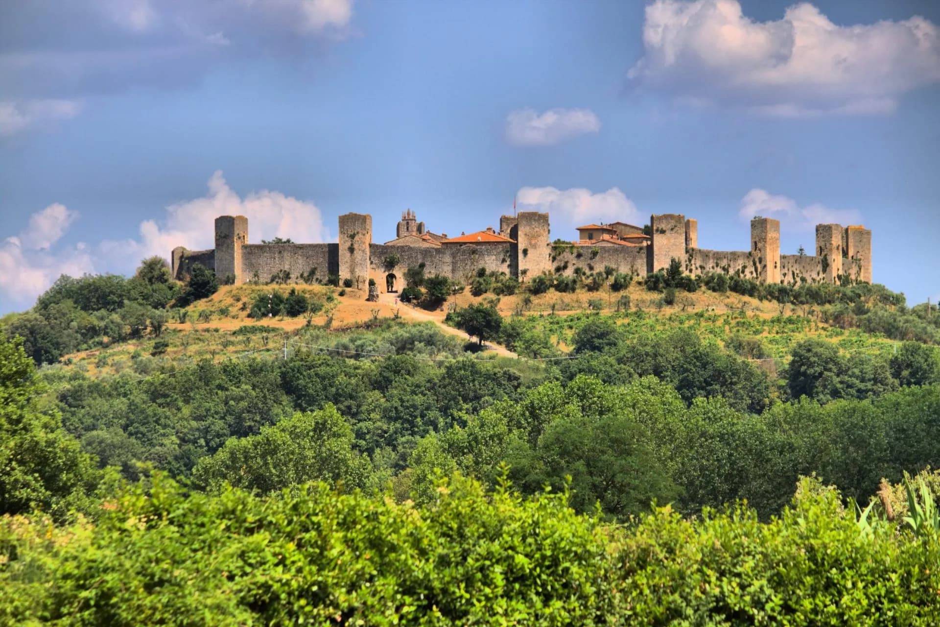 Fortified medieval walls of Monteriggioni on a hill above lush green trees under a blue sky.