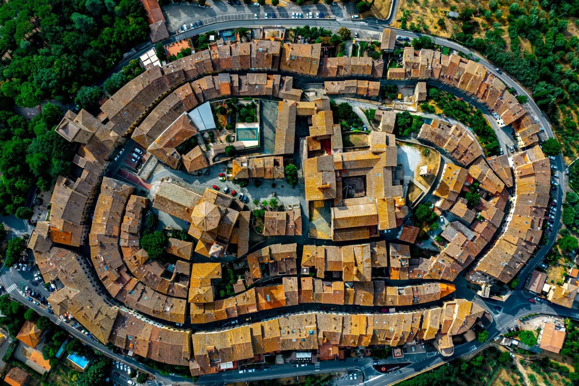 Aerial view of the circular medieval town walls and terracotta roofs of Lucignano, surrounded by green trees.