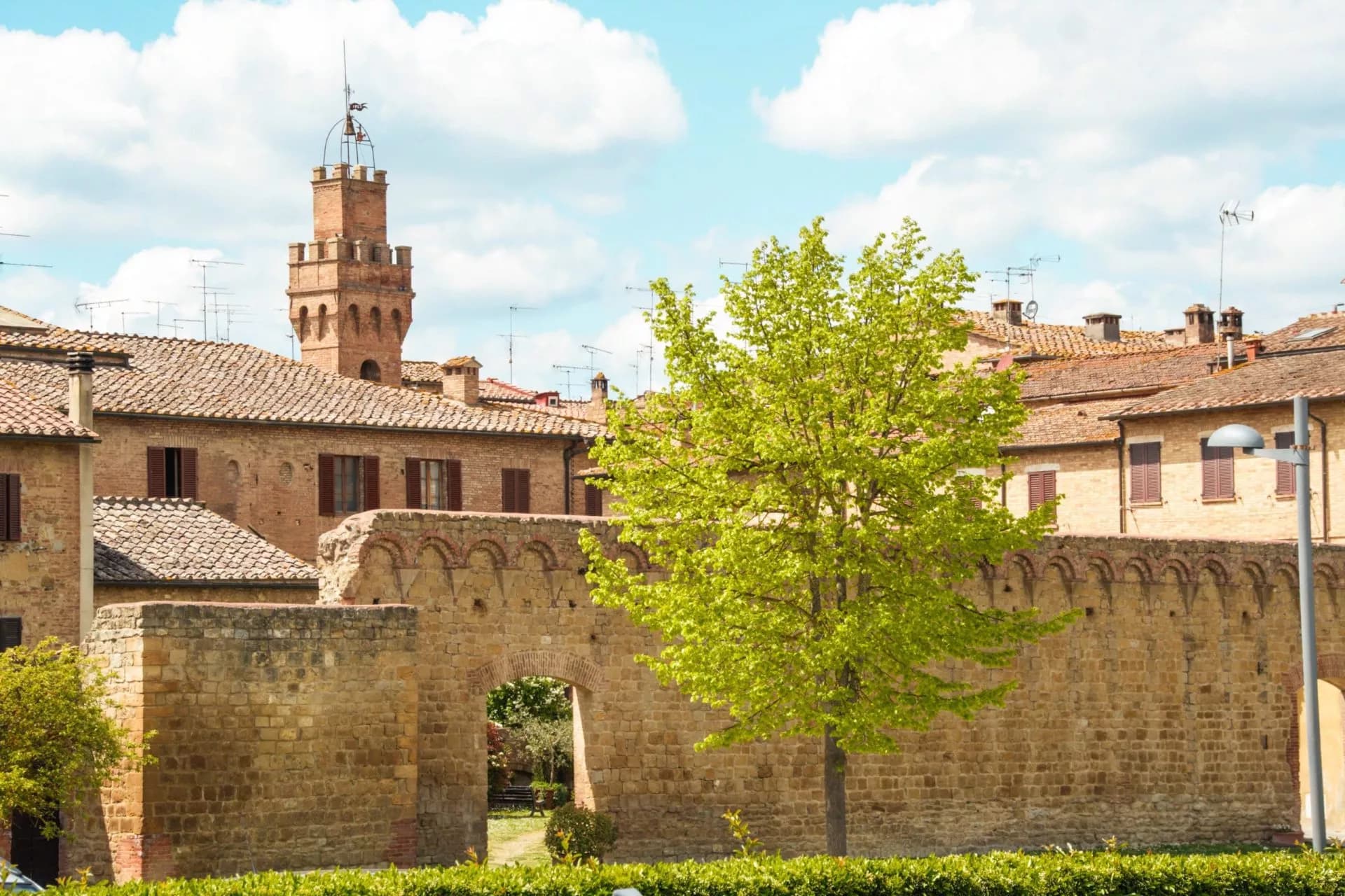 Historic stone wall and tower with terracotta roofs under a blue sky in Buonconvento.
