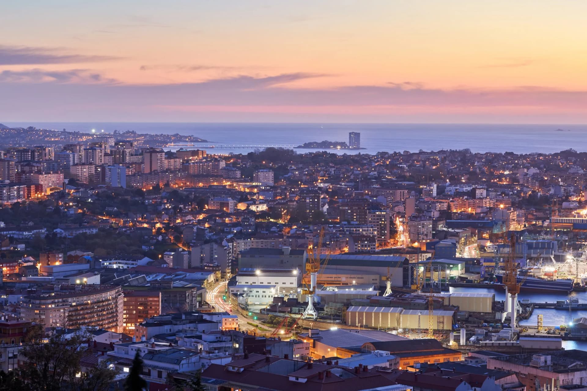 City of Vigo skyline at sunset with illuminated buildings, shipyard, and the sea.