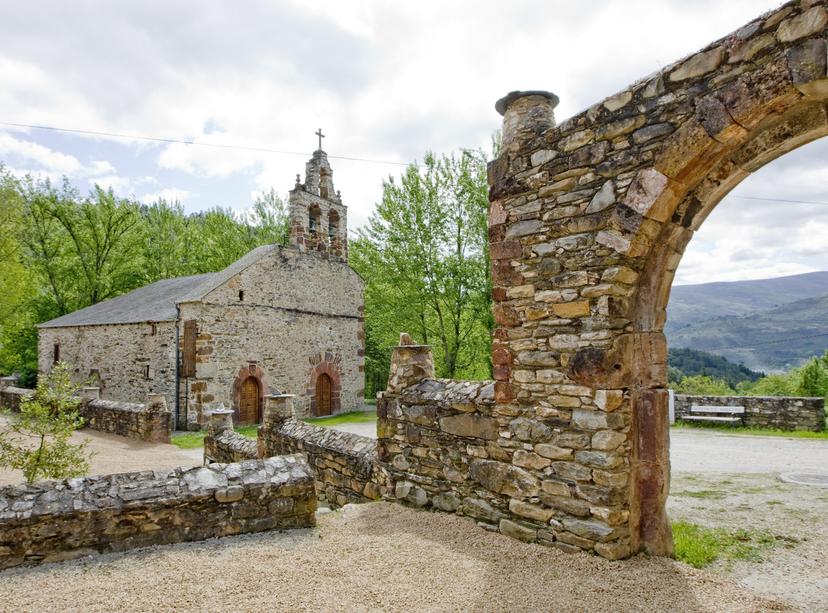 Stone church with bell tower behind a rustic stone archway and wall, green mountains in background.