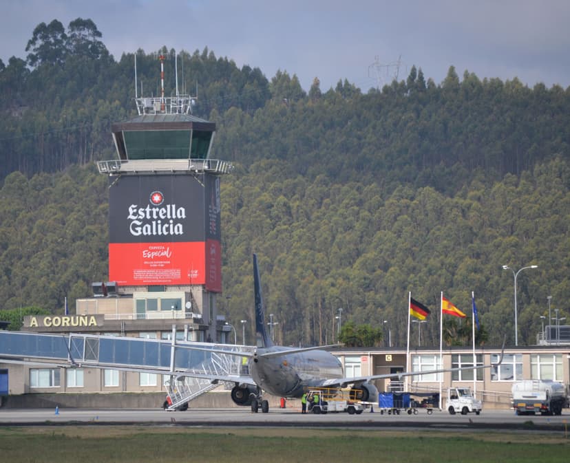Airplane boarding at A Coruña Airport with control tower featuring Estrella Galicia ad and forested hill.