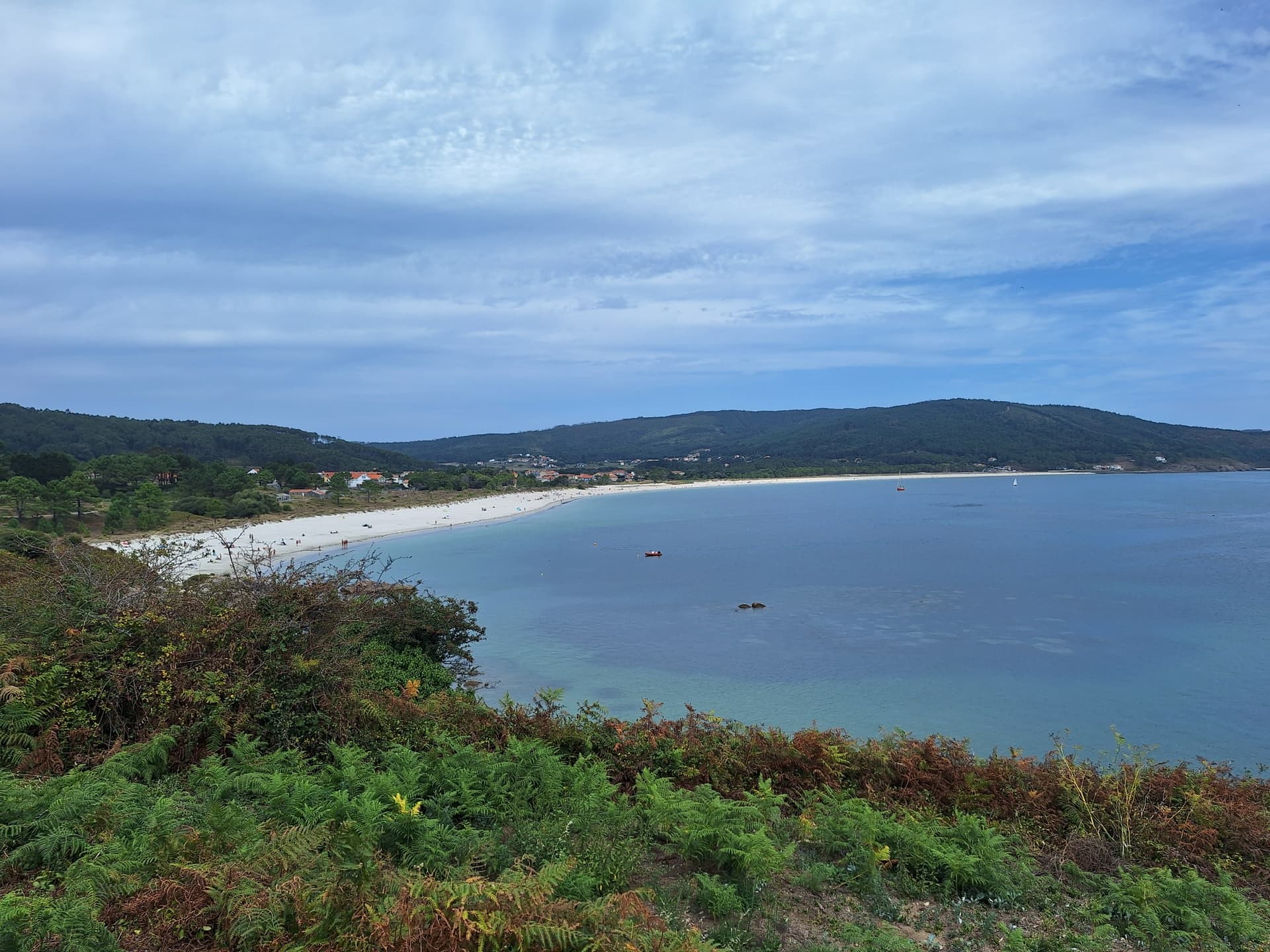 Long sandy beach curving along a bay with calm blue water and forested hills under a cloudy sky