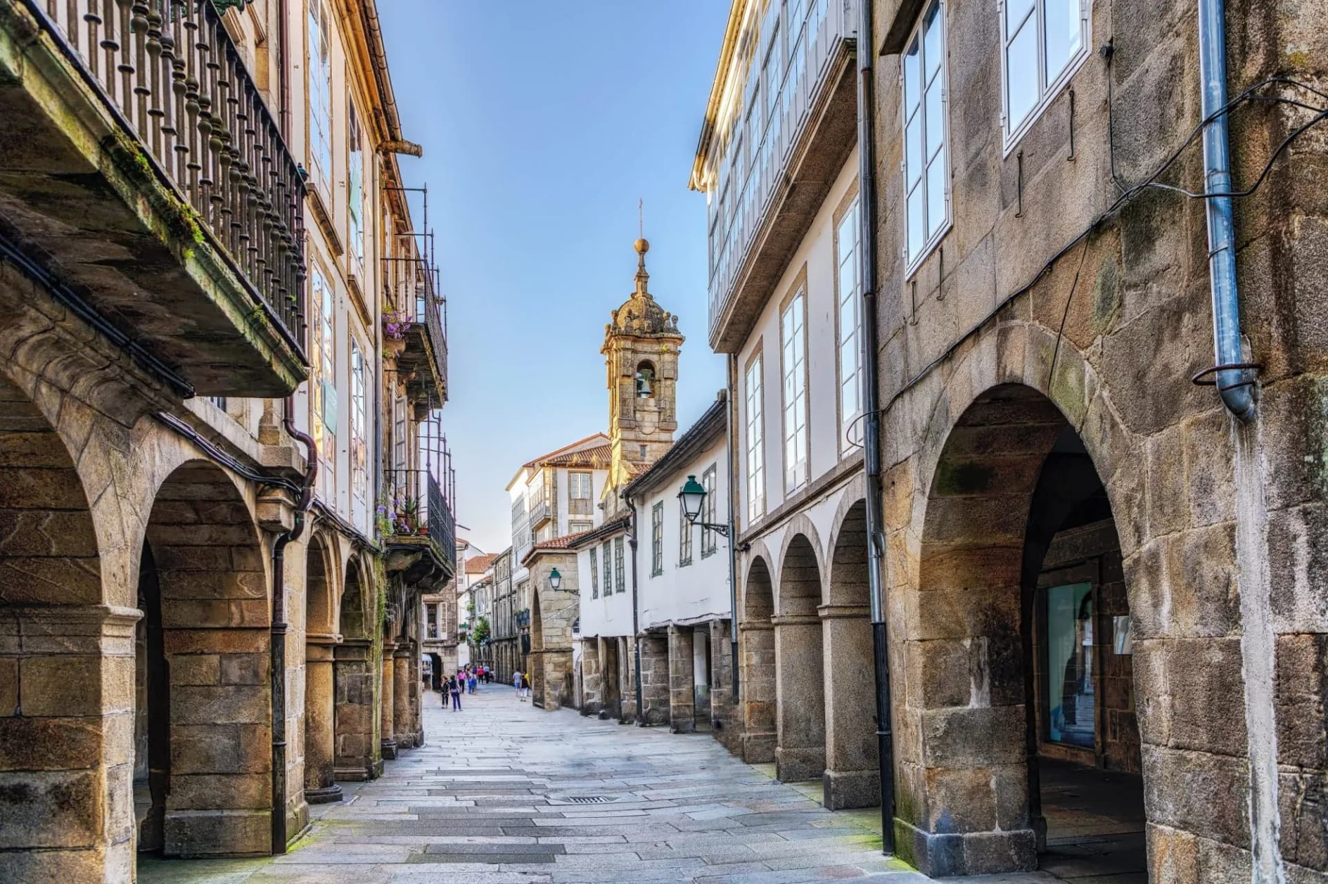 Narrow stone street with arcades leading toward a bell tower in the old town of Santiago de Compostela.