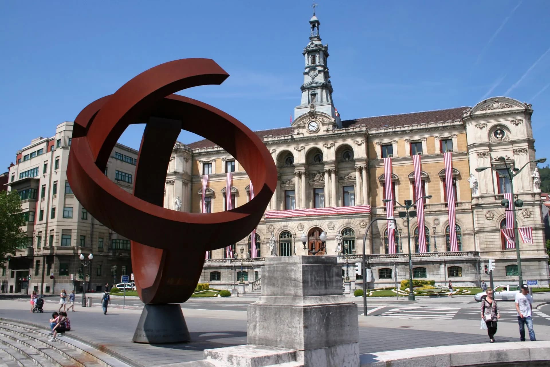 Large abstract sculpture in front of Bilbao Town Hall with flags and clear blue sky.