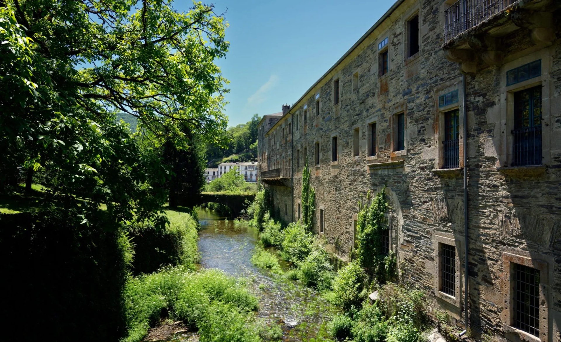 Landmark monastery alongside Sarria river with lush green foliage and clear blue sky.
