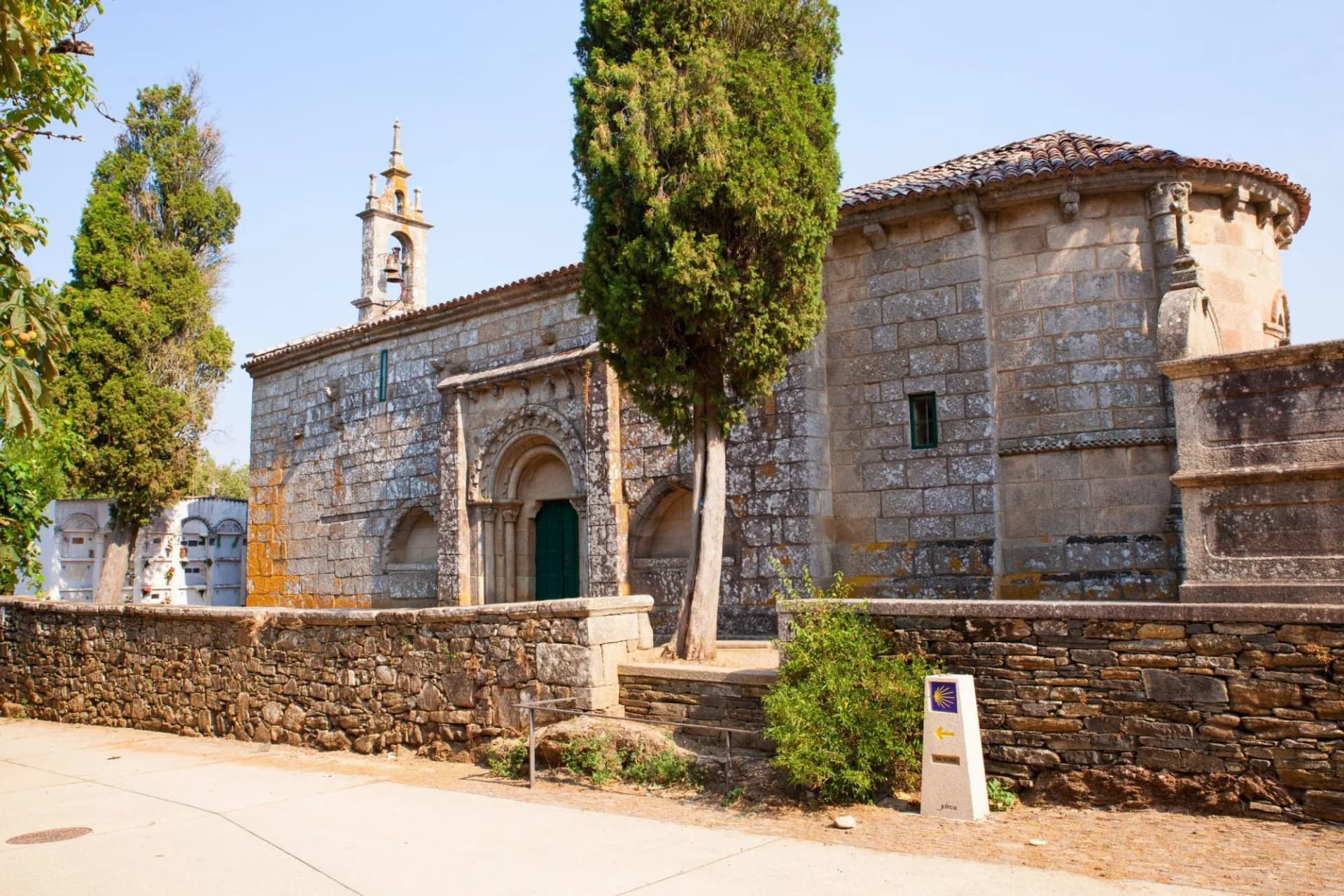 Church of Santa Maria de Melide with stone facade, cypress tree, and Camino de Santiago marker.