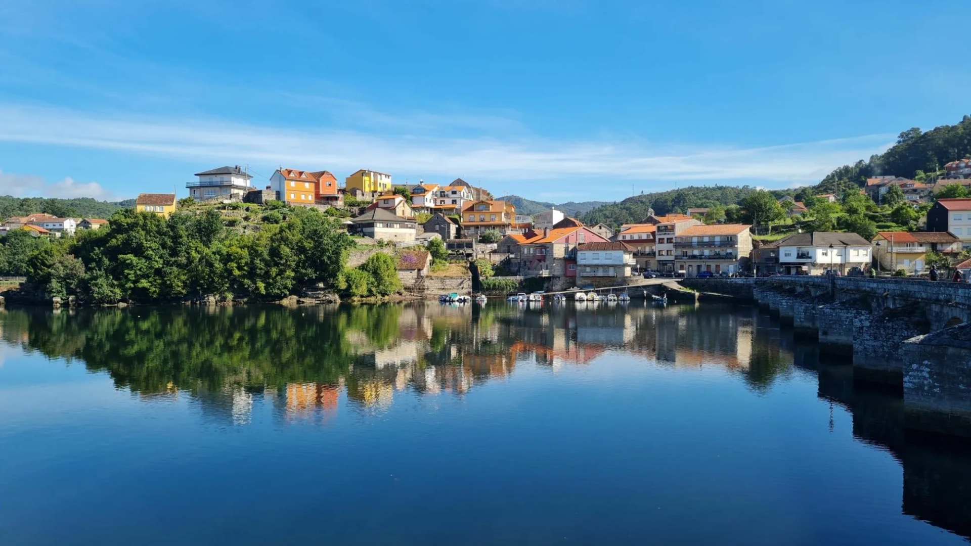 Town on hillside reflected in calm blue water with stone bridge in Caldas de Reis.