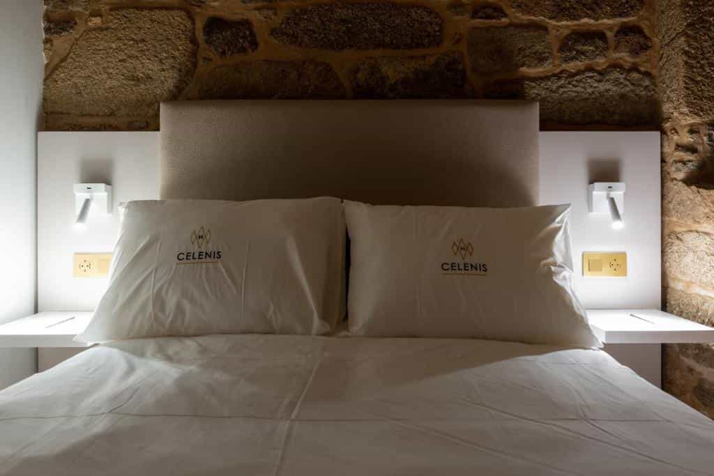 Hotel bed with white linens and pillows featuring "CELENIS" logo against a stone wall.