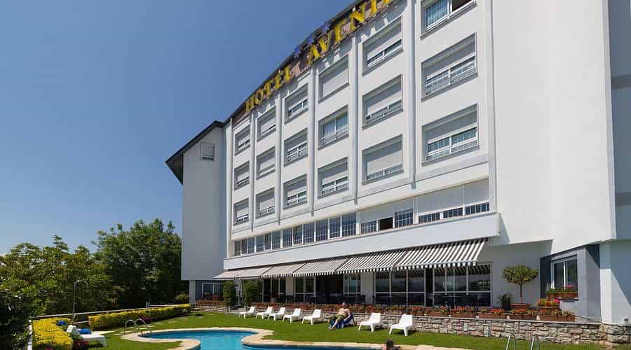 Hotel Avenida exterior with white facade, pool, and sunny green lawn.