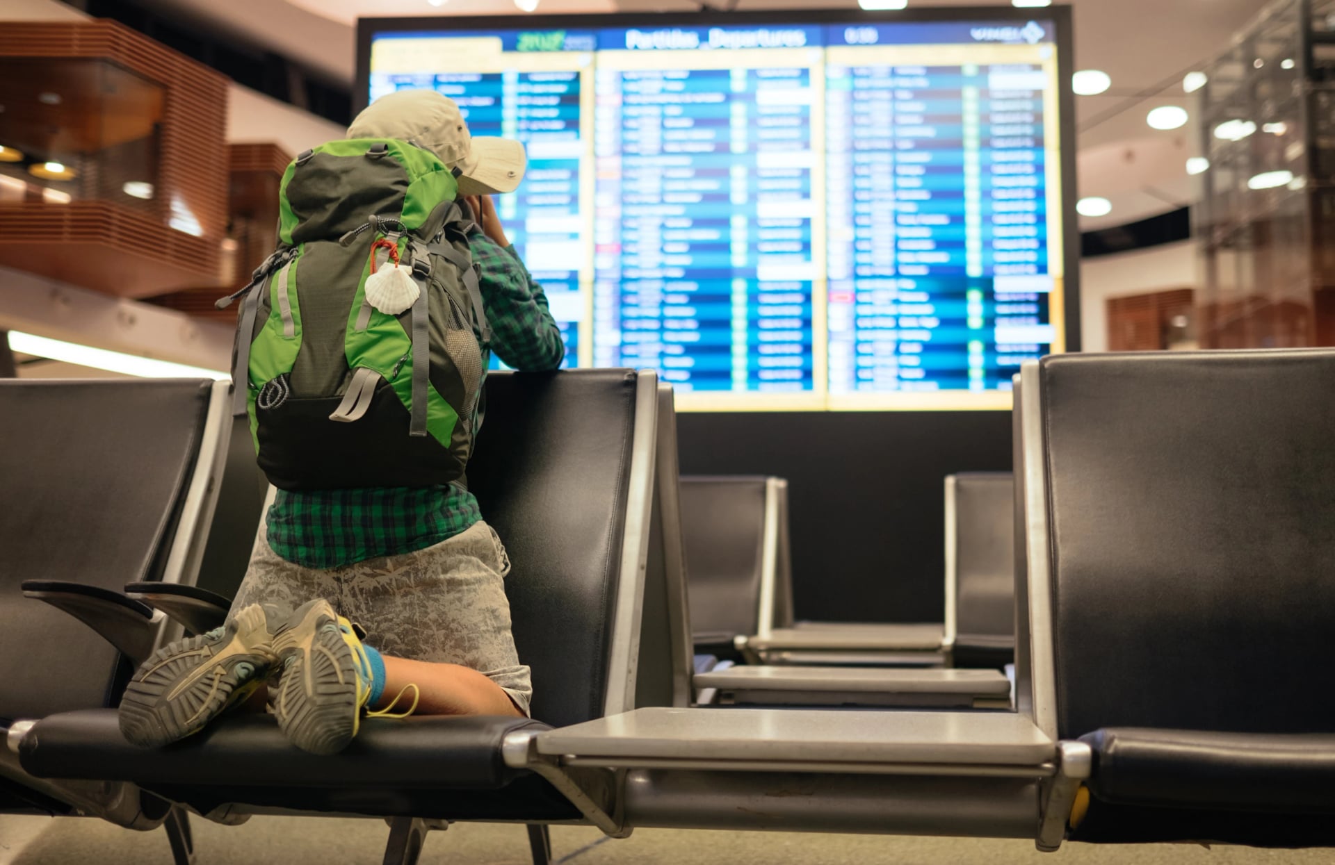 Jovem viajante menino com mochila verde olhando para o painel de partidas do aeroporto. A vibração de aventura captura a antecipação da exploração, planejamento da jornada em um ambiente moderno de terminal. Conceito de viagem e companhias aéreas