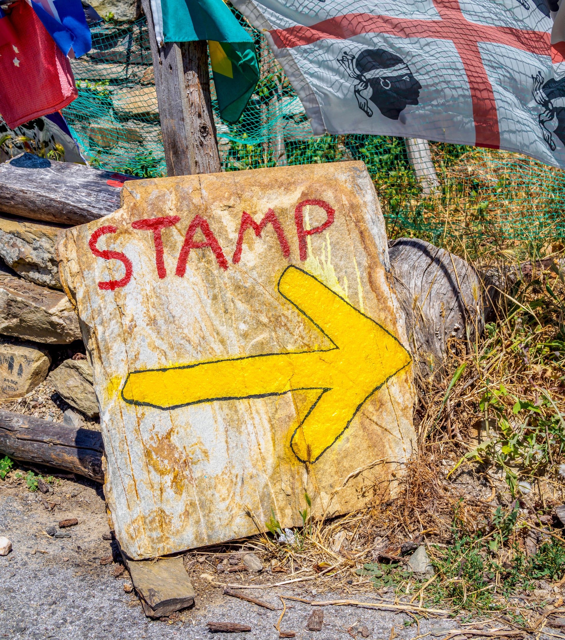 Ein verwittertes Steinschild mit einem gelben Pfeil und dem Wort STAMP, das einen Ort angibt, um das Compostela-Dokument auf dem Camino de Santiago zu stempeln, umgeben von Erinnerungsstücken, die von vorbeiziehenden Pilgern hinterlassen wurden.