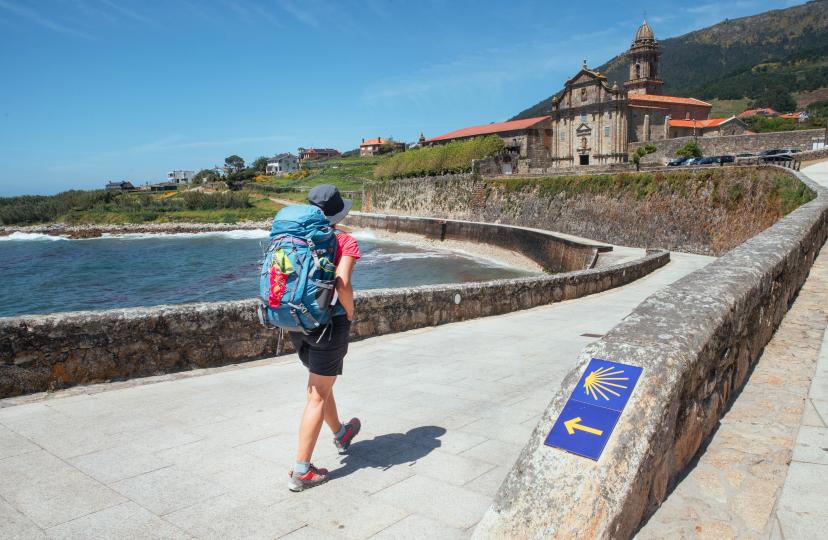 Female pilgrim with backpack walking next yellow arrow and shell directing Camino WAY inspiring solo walk on famous Camino de Santiago, Praia de Oia embankment, Royal Monastery of Santa María de Oia