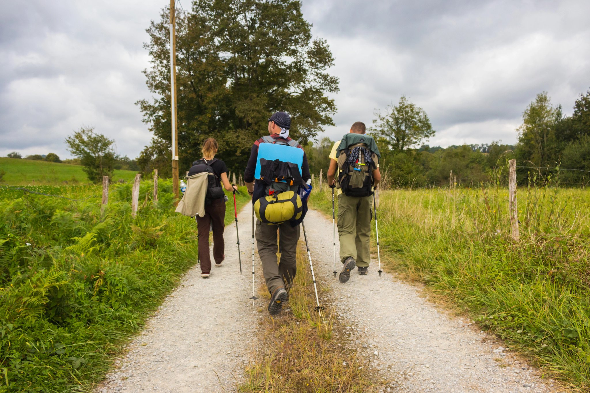 Španielsko - 01/10/2019: Traja neznámi pútnici kráčajúci po Camino de Santiago. Skupina turistov s batohmi na dedinskej ceste. Aktívni ľudia na ceste. Pútnická koncepcia. Turisti zozadu.