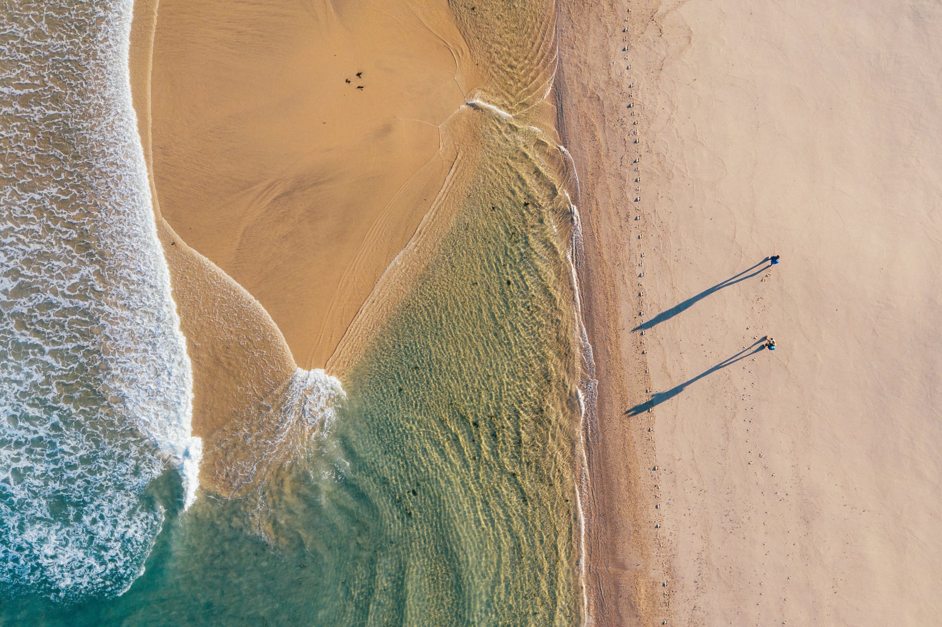 Vogelperspektive auf ein Paar, das entlang eines Sandstrandes geht, der von den Wellen des Atlantischen Ozeans in Portugal gewaschen wird und lange Schatten im schwachen Licht der Morgensonne wirft. Aufnahme während des Camino Portugués – beliebte Pilgerroute