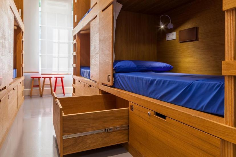 Wooden hostel bunk beds with blue bedding, open storage drawer, and bright window light.