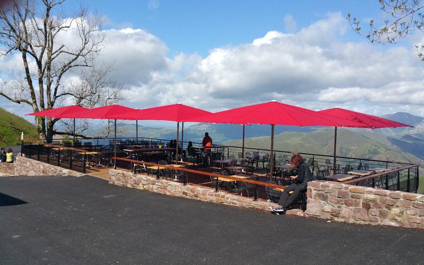 Outdoor terrace with red umbrellas overlooking green mountains under a cloudy blue sky.
