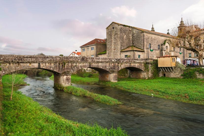 Puente de Santiago over the Sar River in Padrón, Pontevedra, surrounded by lush greenery, historic architecture, and rustic charm under a soft cloudy sky in rural Spain..