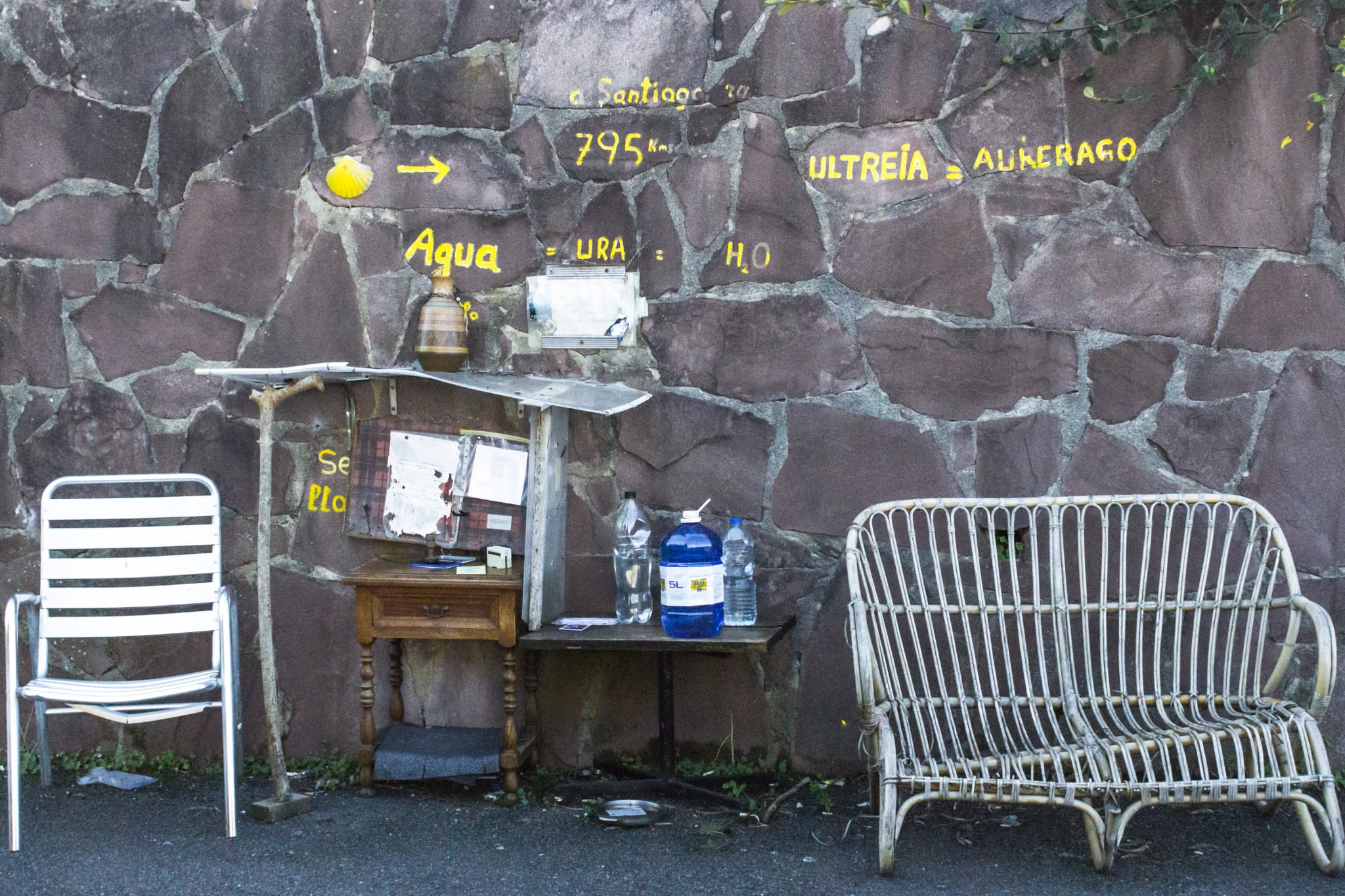 Camino de Santiago. Droga św. Jakuba. Santiago de Compostela. Miejsce do relaksu dla pielgrzymów. Darmowa woda dla podróżnika. Meble ogrodowe. Żółte napisy jako wskaźnik trasy.