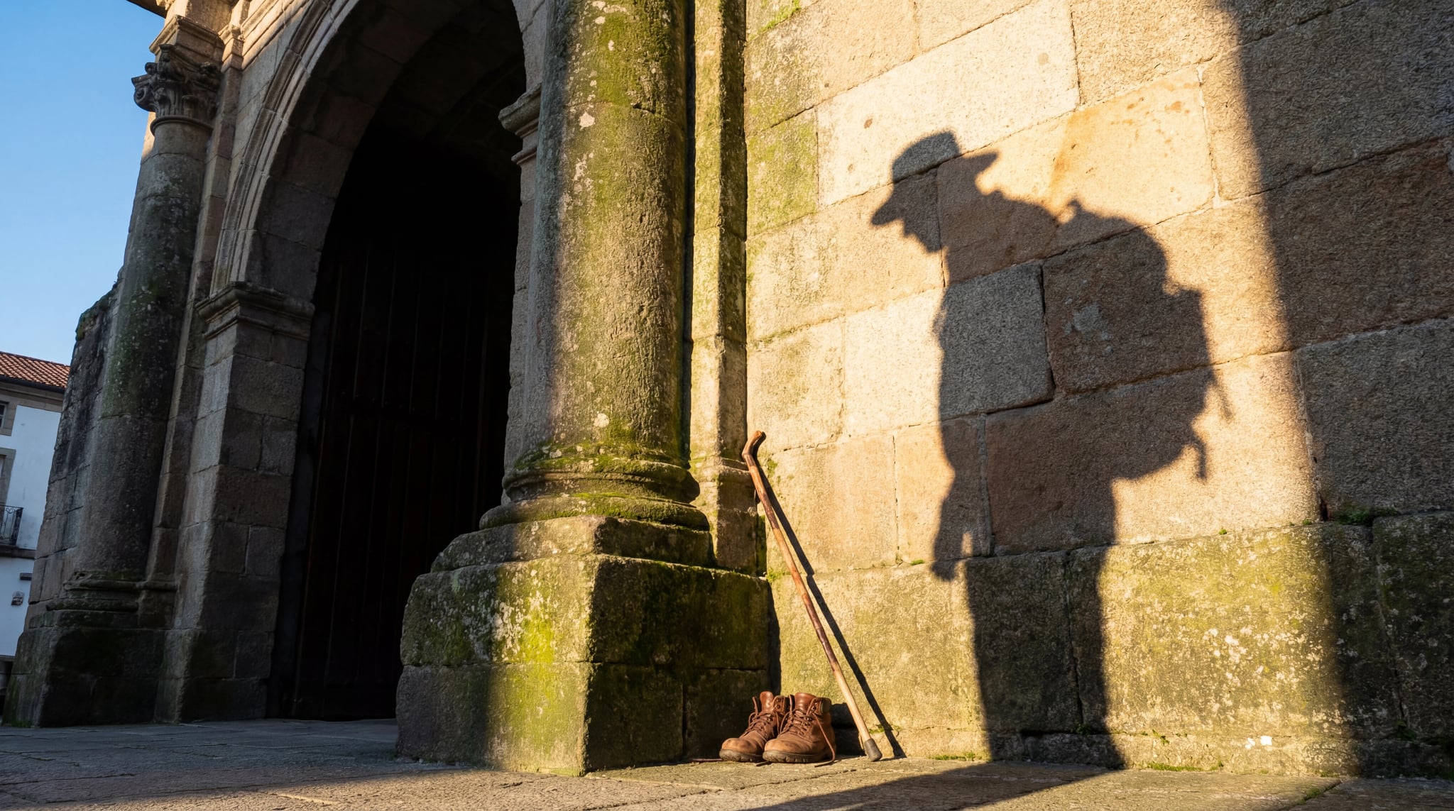 De schaduw van een vermoeide reiziger valt op de oude stenen muur van een kerk in Spanje. Laarzen en een wandelstok rusten in de zon, wat een pauze op de Camino de Santiago suggereert.