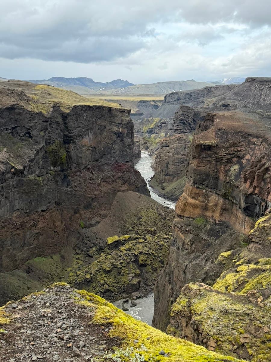 River flowing through a deep volcanic canyon with mossy rocks and distant mountains under a cloudy sky