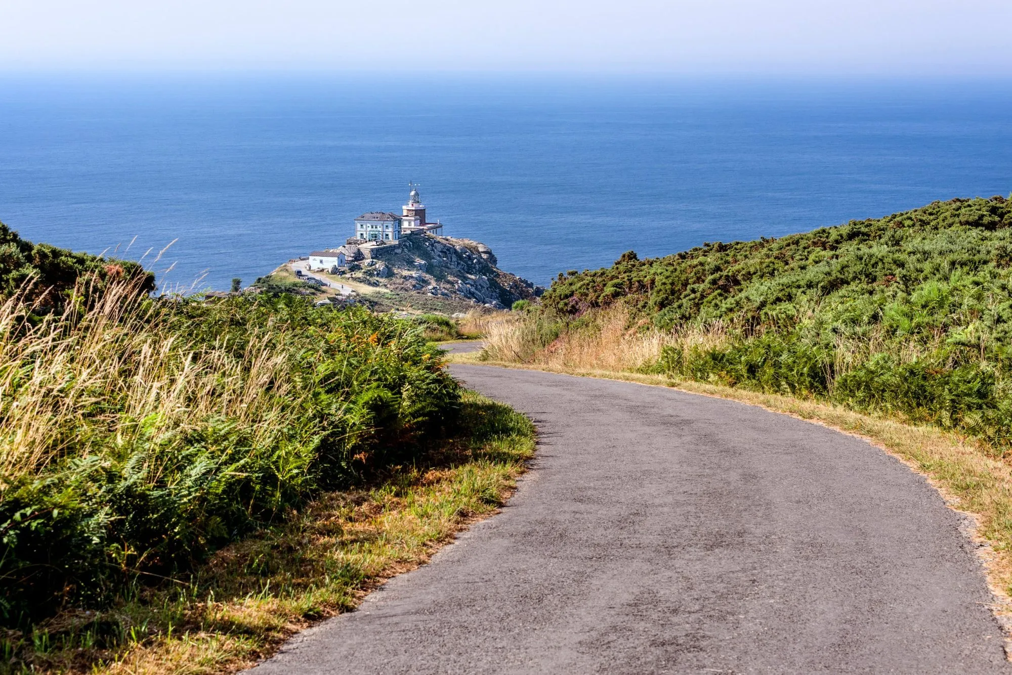 Spain, Finisterre: Famous lighthouse on rocky cliff with road, seascape, ocean sea, skyline and blue sky in the background. It is located on a rock-bound peninsula and was known as end of the world.