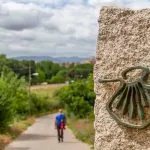 Pilgrim out of focus walks along the Camino de Santiago (Sant James Way) carrying a backpack next to a column on focus with the shell symbol of the Camino Santiago de Compostela